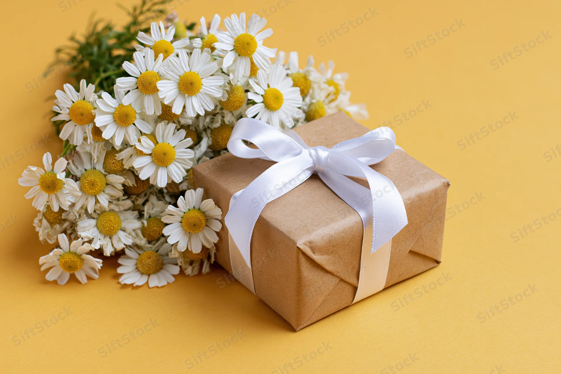 Kraft paper gift box tied with white satin ribbon bow beside a bouquet of daisies on yellow background
