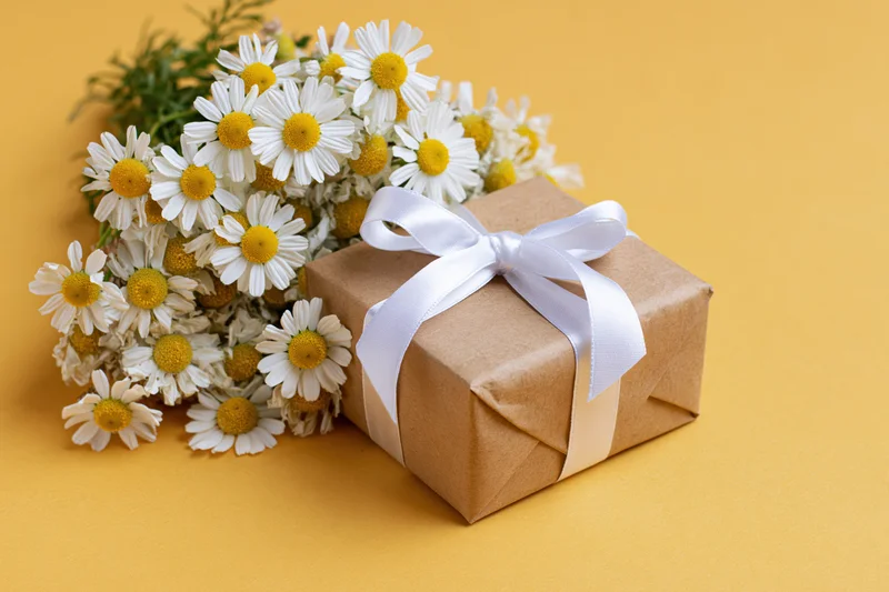 Kraft paper gift box tied with white satin ribbon bow beside a bouquet of daisies on yellow background