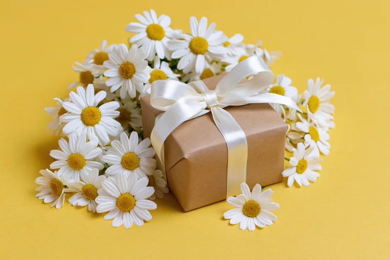 Small kraft paper gift box tied with white satin ribbon bow surrounded by daisies on yellow surface