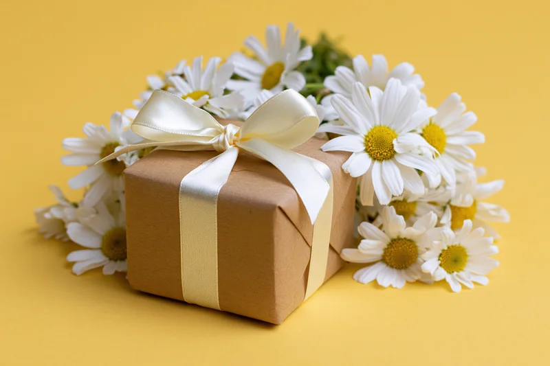 Kraft paper gift box tied with white satin ribbon bow surrounded by daisies on yellow background
