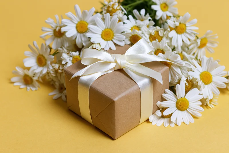 Kraft paper gift box tied with white satin ribbon bow surrounded by daisies on yellow background