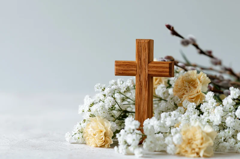Wooden cross standing among white gypsophila and beige carnations on a light surface