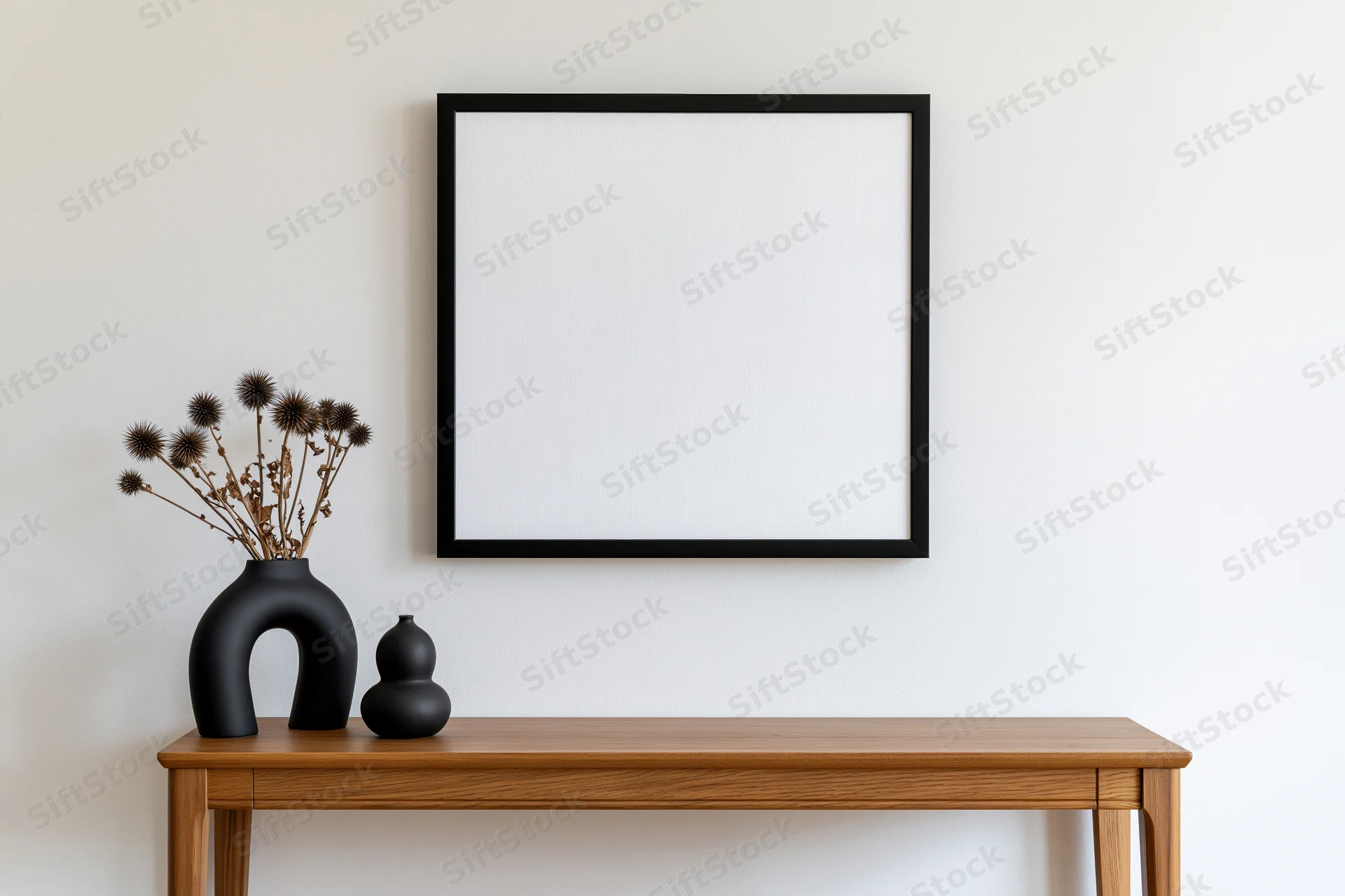 Square black-framed empty white canvas mounted on pale white wall above wooden console table with black vases and dried flowers