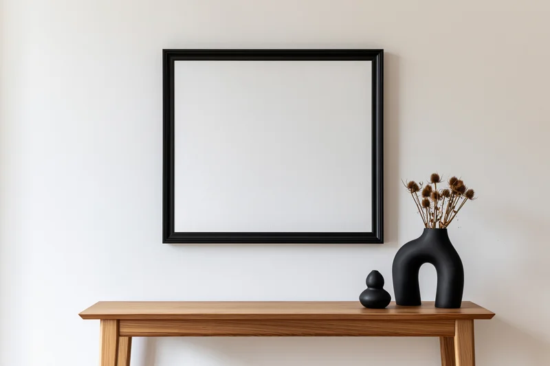 Square black frame mounted on wall above wooden console with black vase holding dried flowers