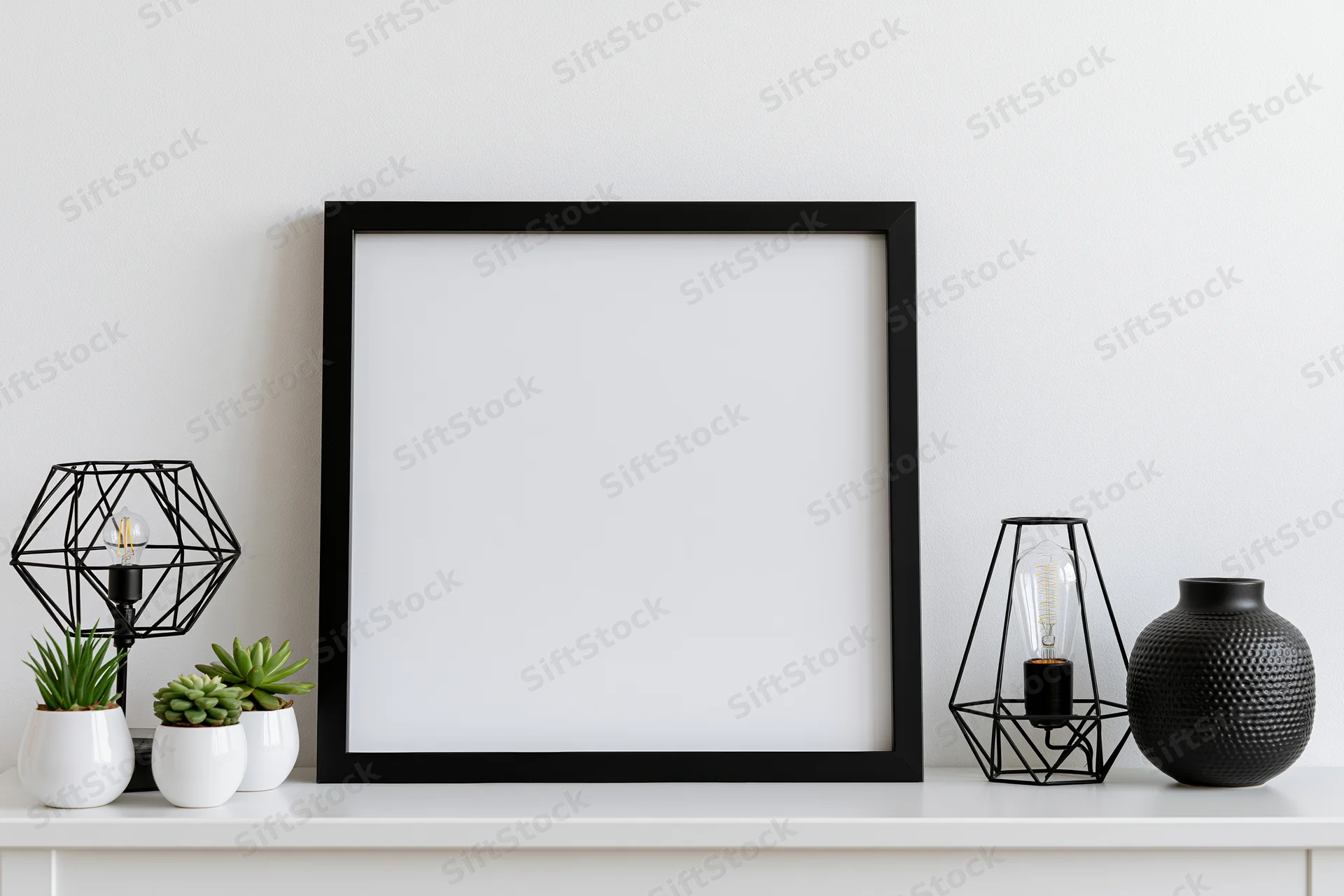 Square black frame with blank white interior on white shelf surrounded by decorative objects including lamps, succulents and a textured vase