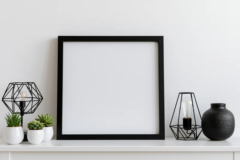Square black frame with blank white interior on white shelf surrounded by decorative objects including lamps, succulents and a textured vase
