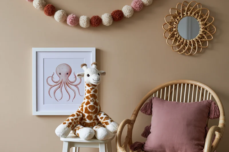 Stuffed giraffe toy sitting on a white stool beside a rattan chair with a pink tasseled pillow, under a framed octopus print and wall decor in a children's room