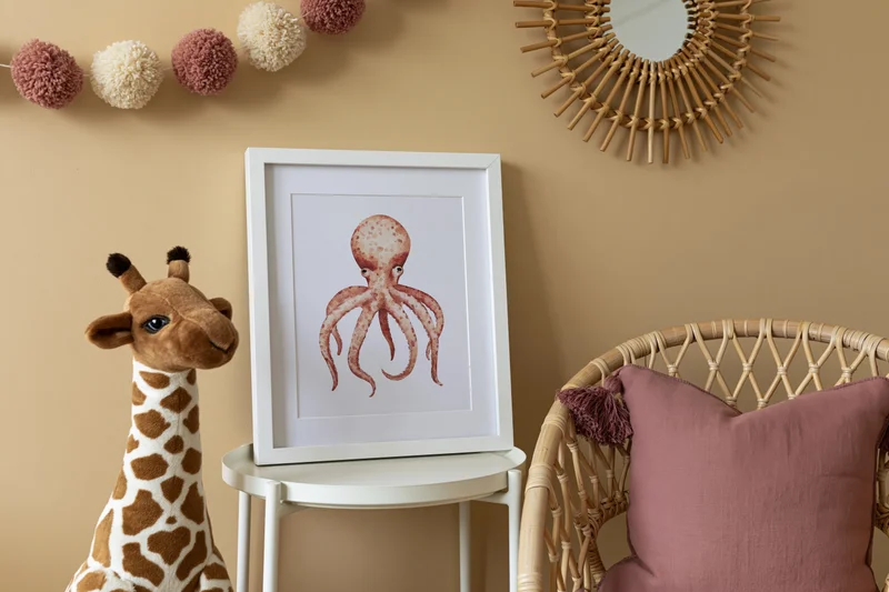 Framed octopus print on a white side table beside a plush giraffe and rattan chair in a children's room