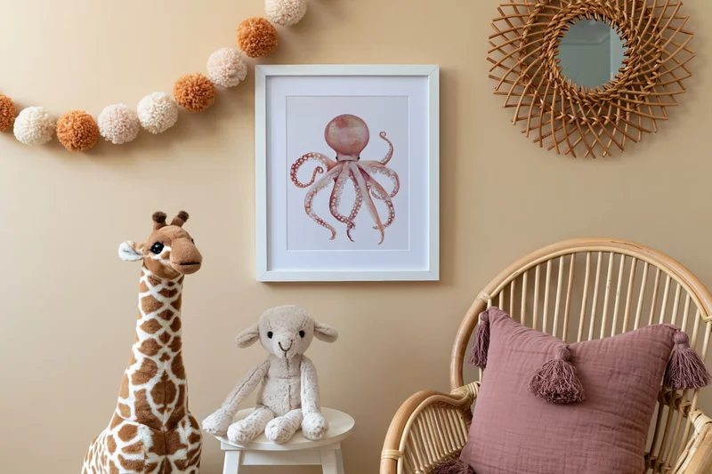 Framed octopus artwork on beige wall above stuffed giraffe and lamb in childrens room with rattan chair and pompon garland