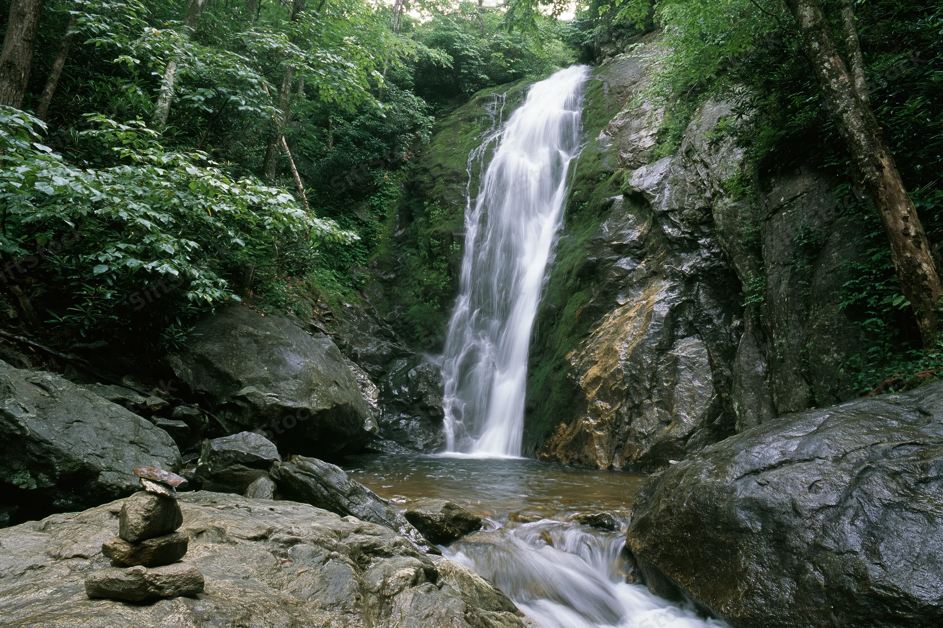 Waterfall cascading down moss-covered rocks in a dense forest surrounded by trees and boulders