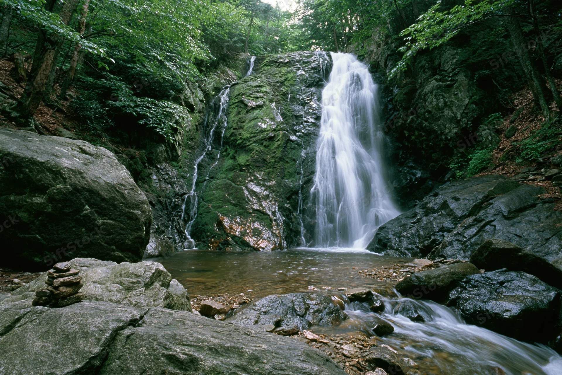 Waterfall cascading down moss-covered rocks in a dense forest with a stream flowing below