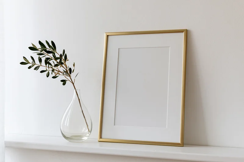 Gold frame leaning against white wall on shelf beside glass vase with olive branch