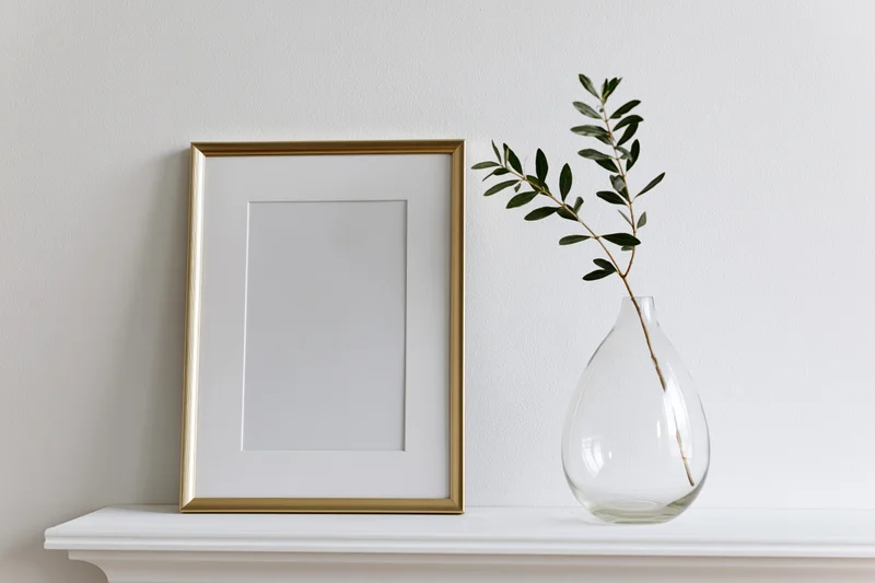 Gold picture frame with blank white mat and olive branch in glass vase on white shelf against wall