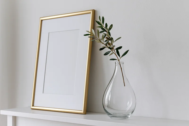 Gold picture frame leaning on white shelf beside glass vase with olive branch