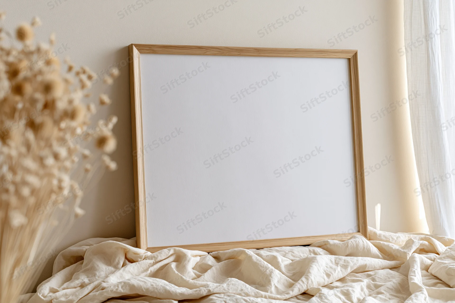 Wooden picture frame leaning against wall on rumpled bedding near dried flowers by window