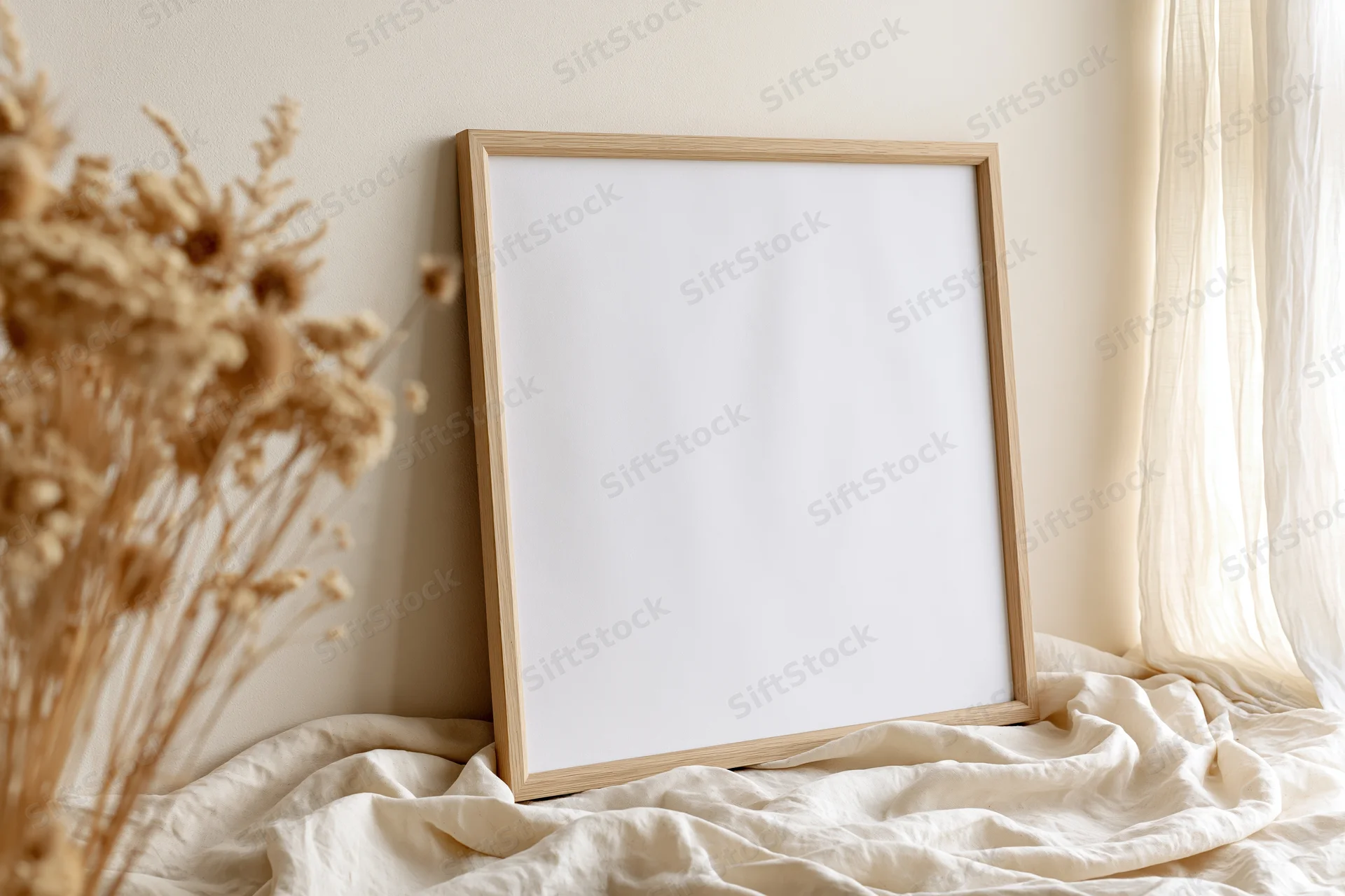 Wooden picture frame leaning against wall on rumpled linen bedsheet near dried flowers and sheer curtain