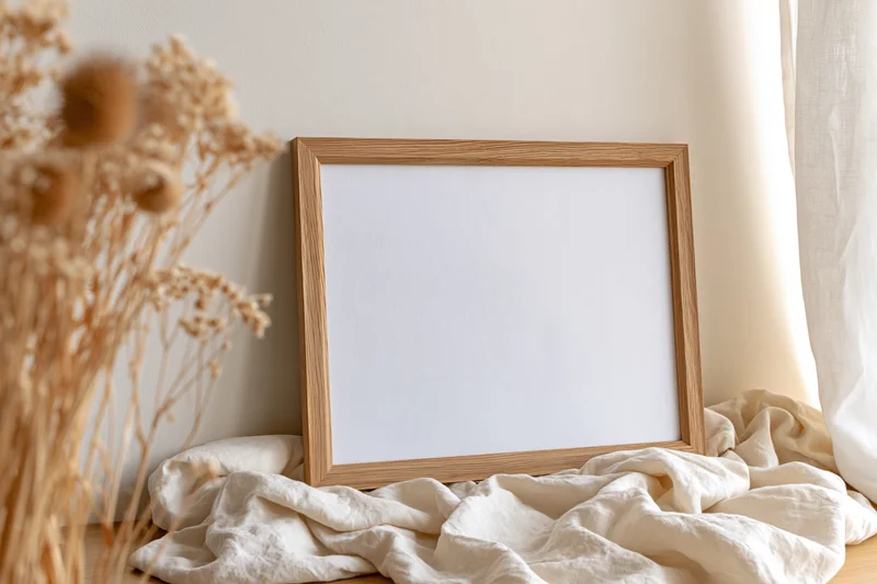 Wooden picture frame leaning against wall on draped fabric near dried flowers by window
