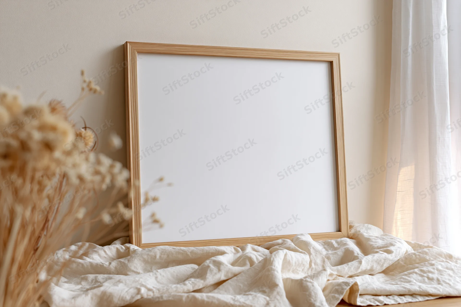Wooden picture frame leaning against wall on crumpled fabric near window with dried flowers