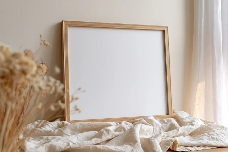 Wooden picture frame leaning against wall on crumpled fabric near window with dried flowers