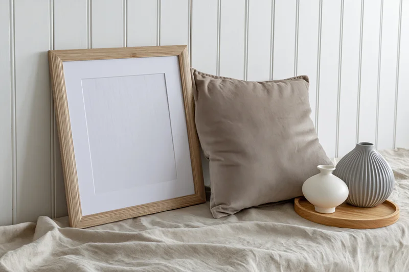 Wooden picture frame leaning against white beadboard wall beside beige pillow and ceramic vases on round tray on bed