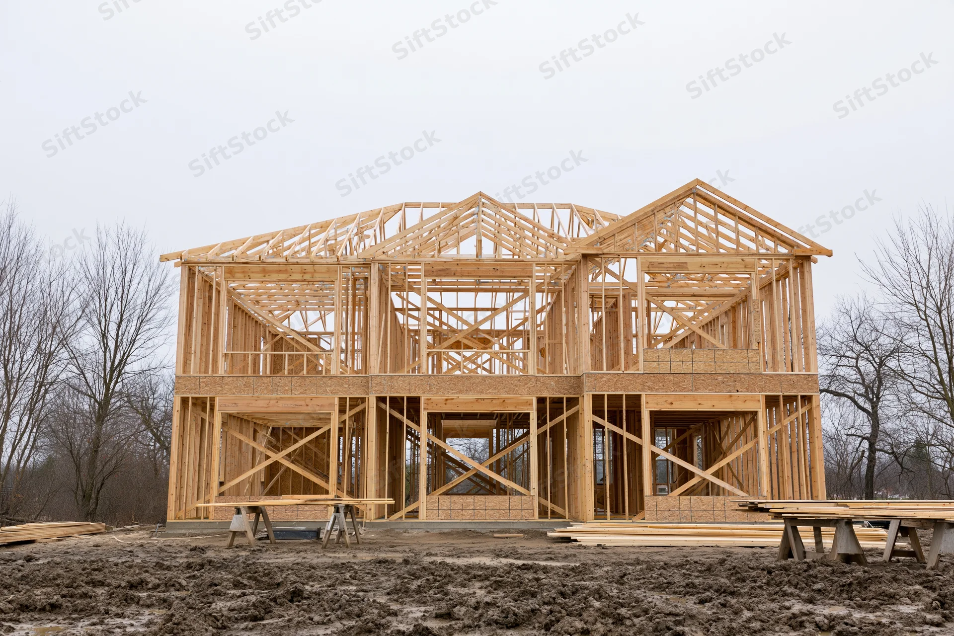 Two-story residential house under construction with exposed wooden framing on a muddy site
