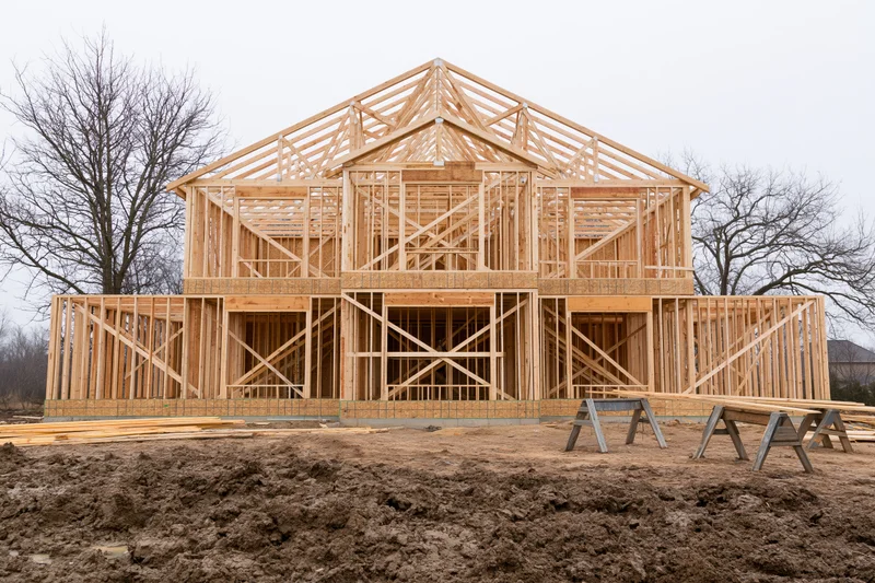 Two-story residential house under construction with wooden frame exposed on muddy site