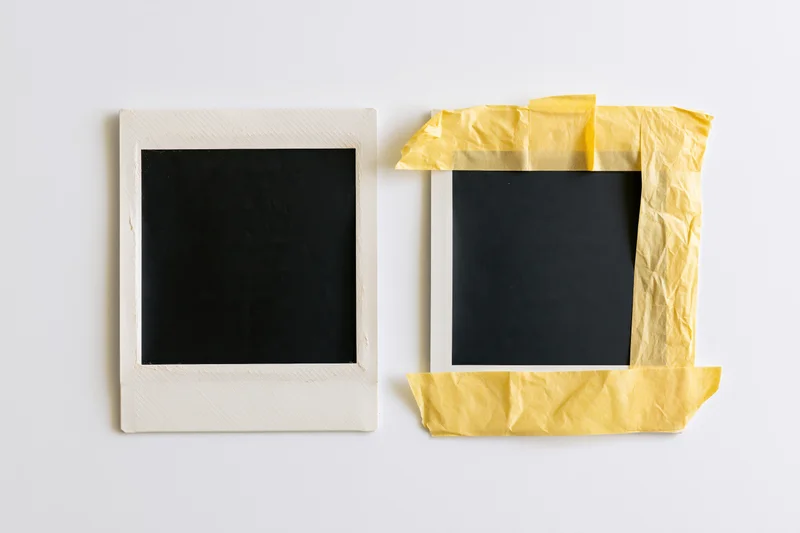 Two blank instant film photo prints side by side on white background, one with yellow tape border