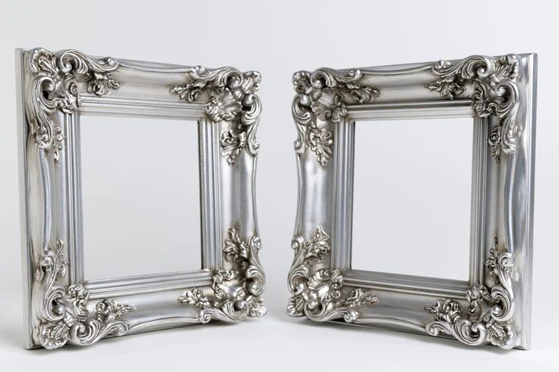 Two ornate silver picture frames with baroque and rococo carvings standing side by side on a white surface