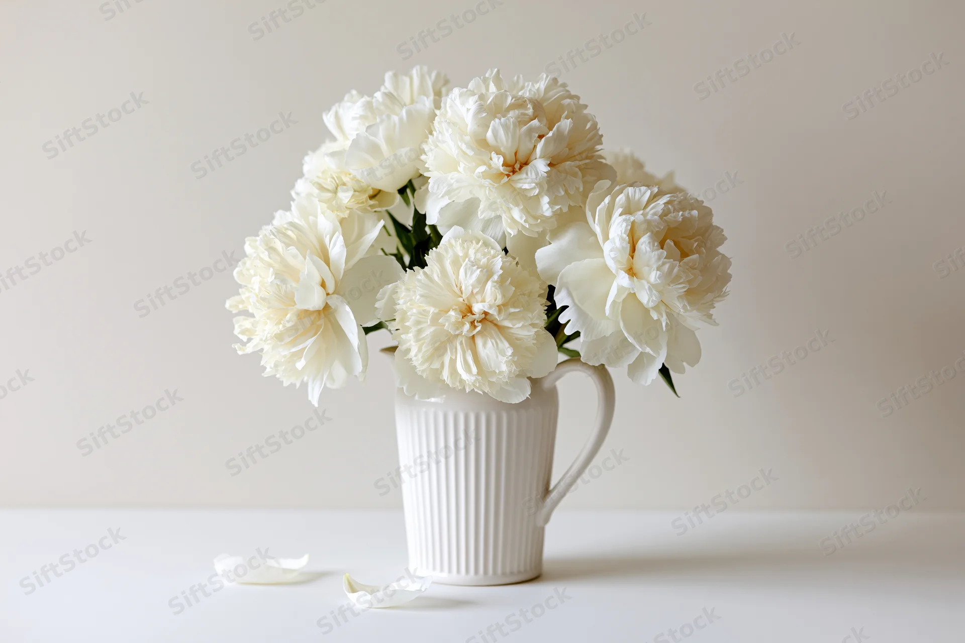 White peony bouquet in ribbed ceramic pitcher on white surface with fallen petals