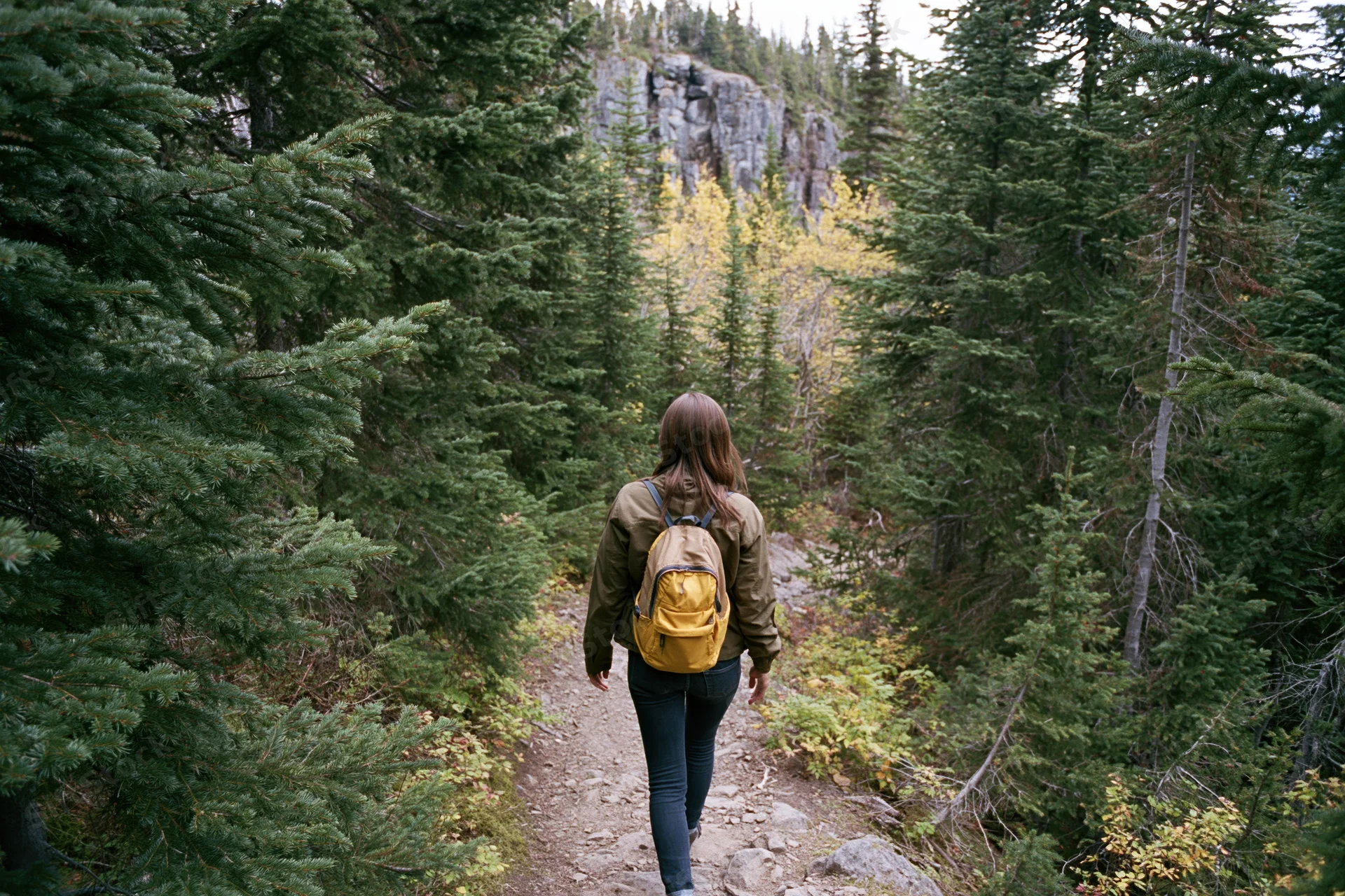 Woman with yellow backpack hiking on forest trail toward rocky cliff