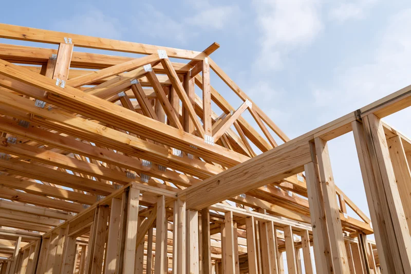 Wooden house frame under construction with exposed lumber beams and trusses against a blue sky