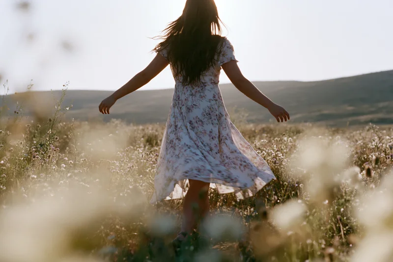 Woman in floral dress with arms outstretched walking through wildflower field at golden hour