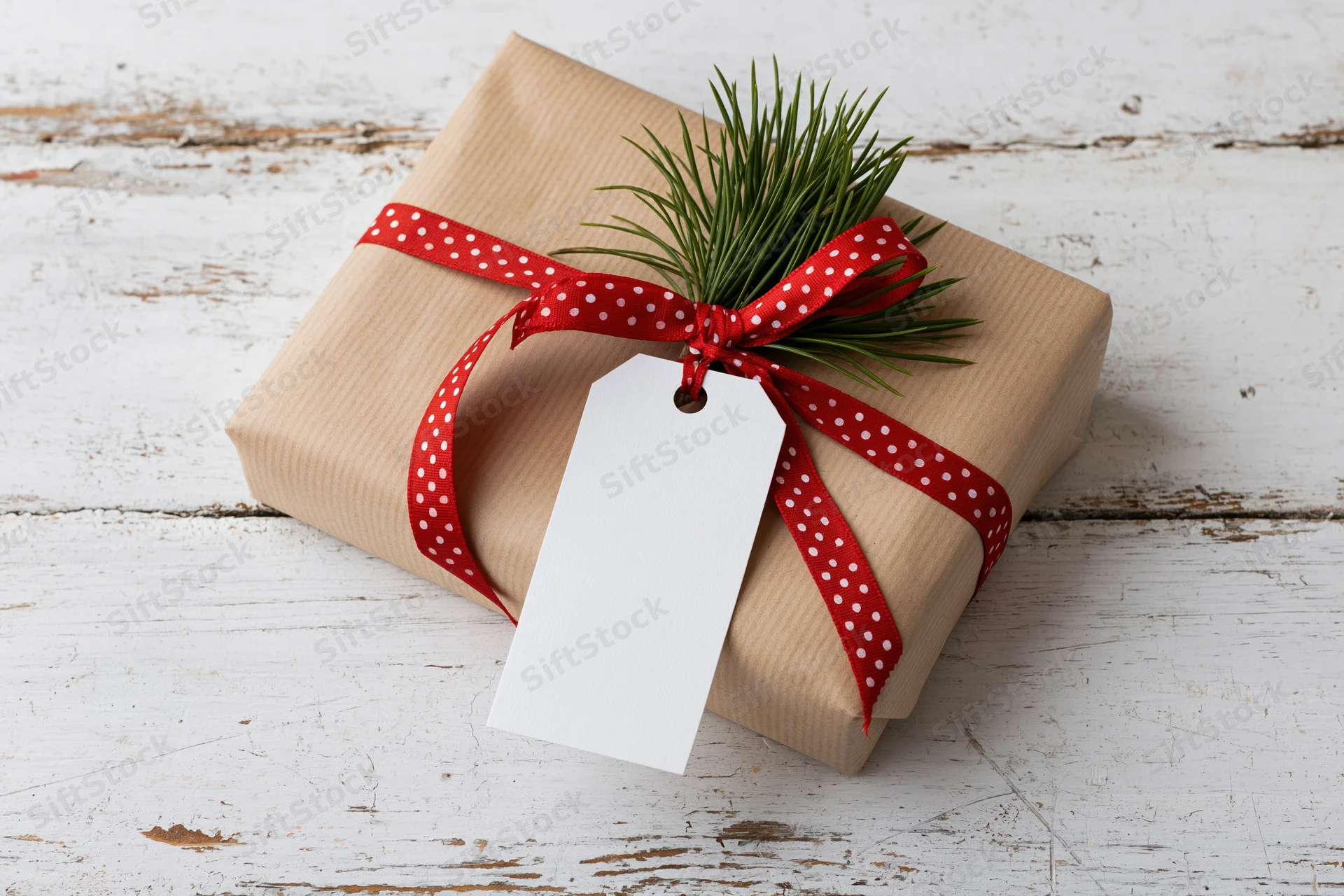 Christmas gift wrapped in brown paper with red polka dot ribbon and pine sprig on white wooden surface