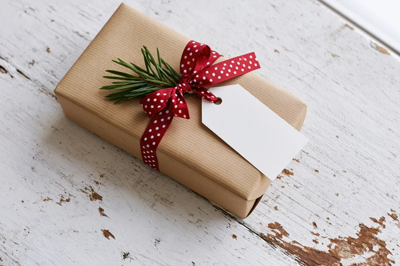Christmas gift wrapped in brown paper with red polka dot ribbon and pine sprig on distressed white wood surface