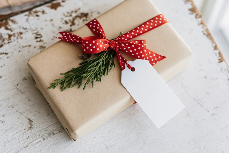 Christmas gift wrapped in kraft paper with red polka dot ribbon and evergreen sprig on white wooden table
