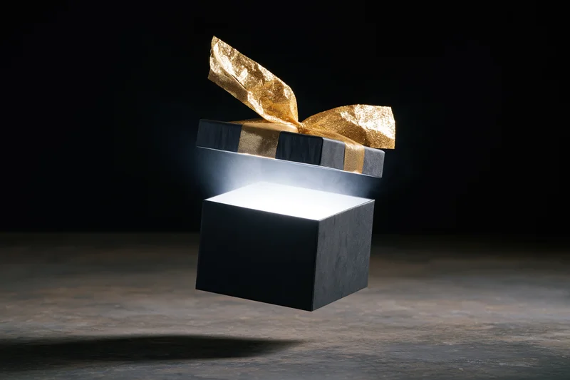 Black gift box with floating lid open and glowing light inside on dark floor