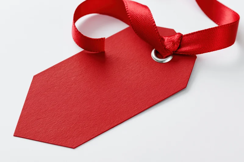 Blank red cardstock gift tag with satin ribbon tied through metal hole on white surface