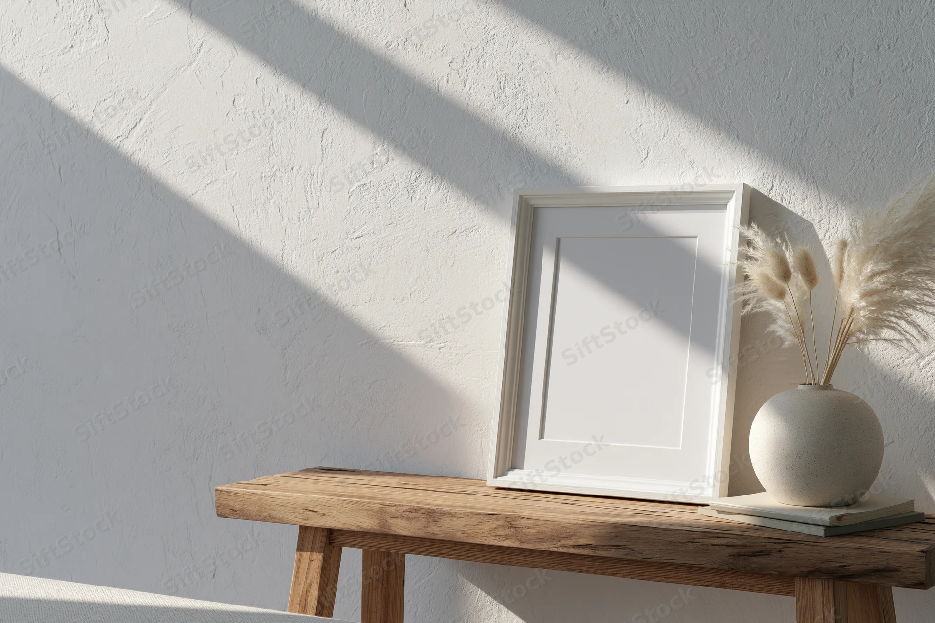 Blank white frame leaning on wooden bench beside vase with pampas grass in sunlit room