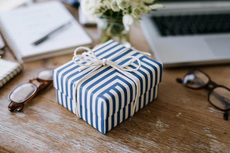 Blue and white striped gift box tied with string on wooden desk with laptop, notebook, and flowers
