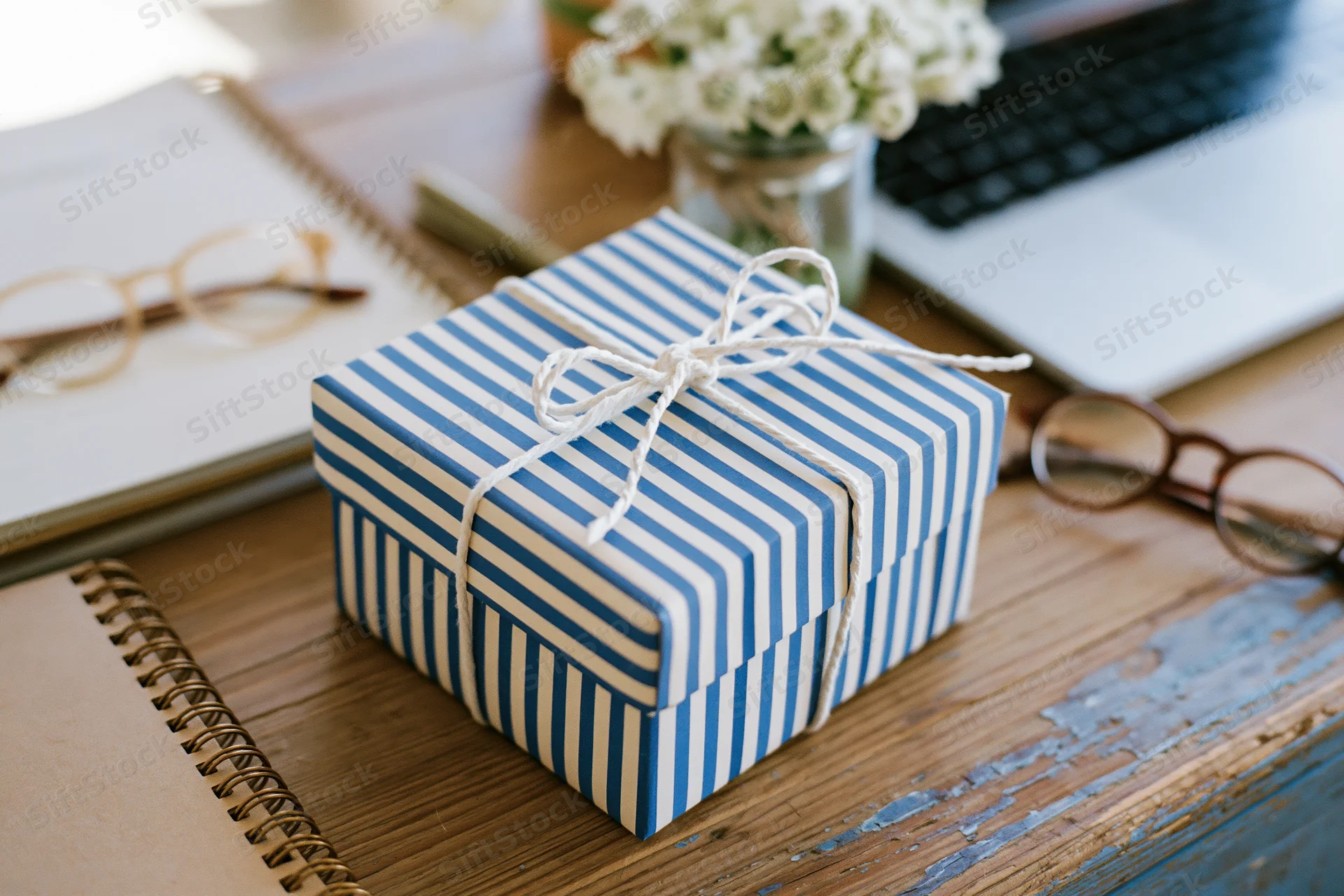 Blue and white striped gift box tied with twine on a wooden desk surrounded by notebooks, glasses, and a laptop
