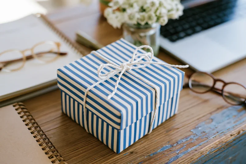 Blue and white striped gift box tied with twine on a wooden desk surrounded by notebooks, glasses, and a laptop