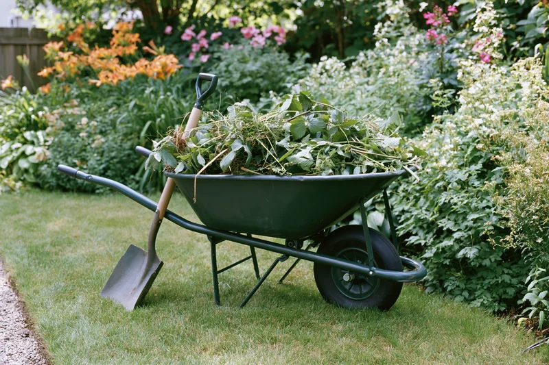 Dark green metal wheelbarrow filled with pulled weeds and a shovel resting beside it on a grassy garden lawn