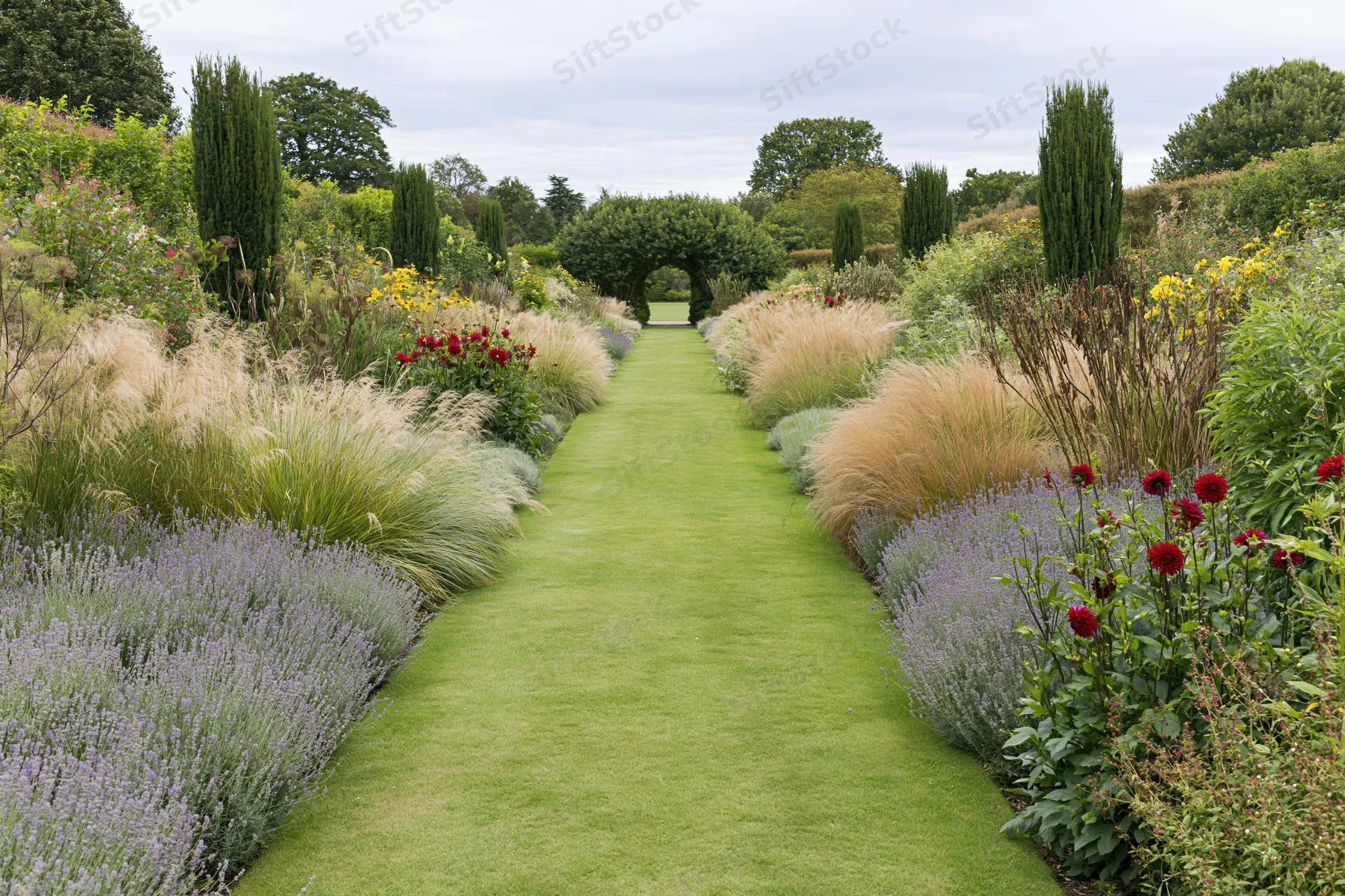 Formal garden path of trimmed green lawn stretching toward a hedge arch, flanked by lavender, roses, and ornamental grasses