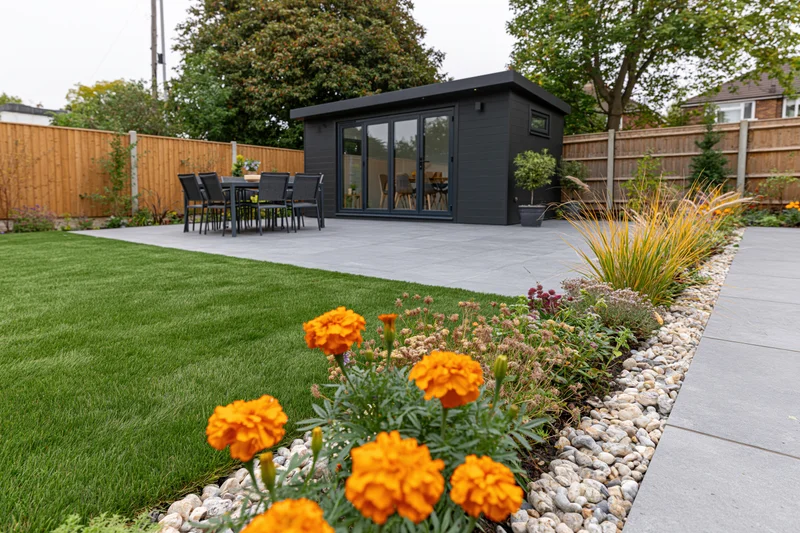 Modern garden with artificial grass, patio, and black shed surrounded by flowers and plants