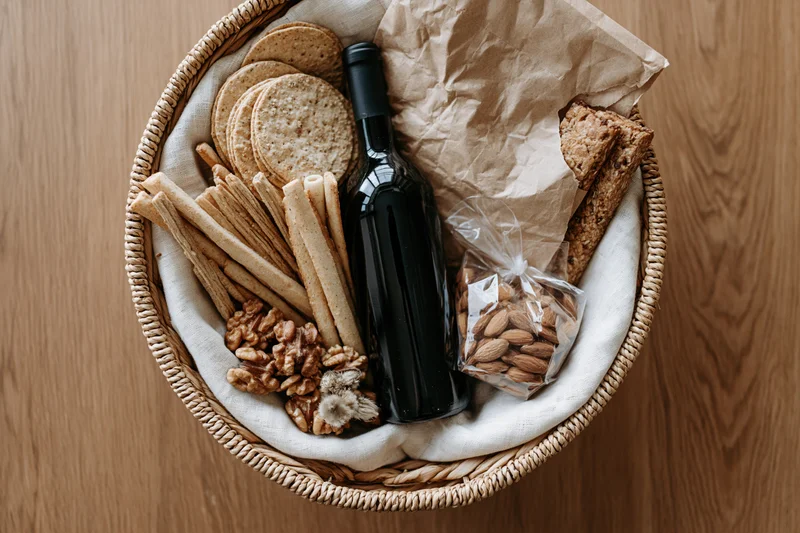 Woven basket filled with dark wine bottle, crackers, bread, almonds, walnuts, and breadsticks on wooden surface