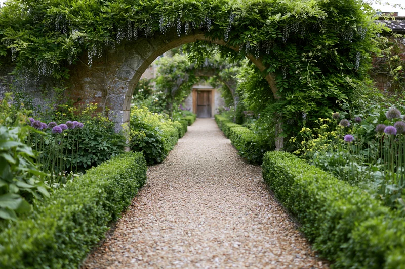 Gravel path leading through stone arches lined with hedges and flowering plants in a formal garden