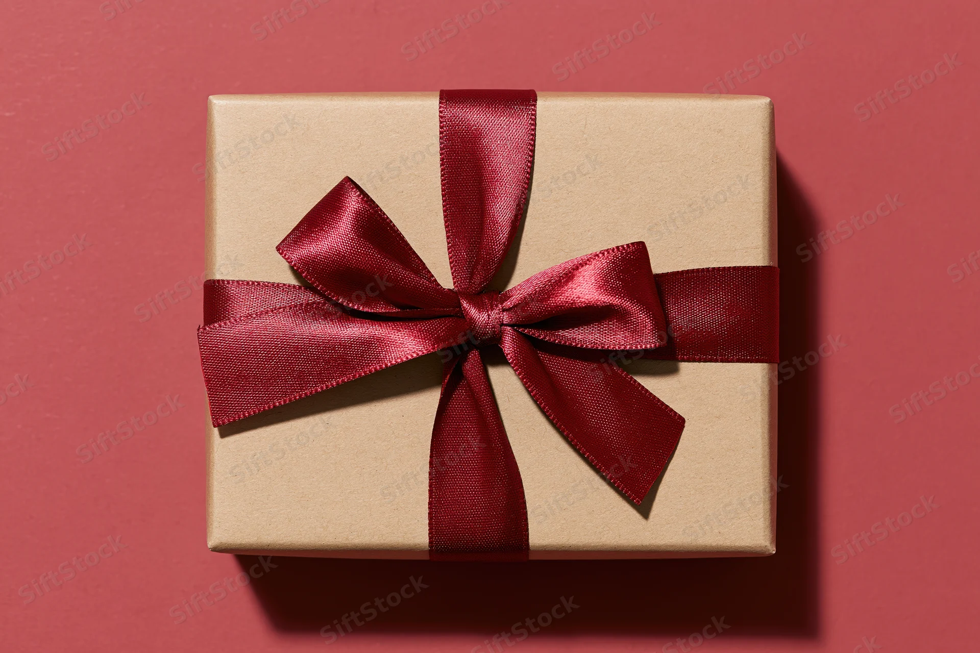 Square kraft paper gift box with red satin ribbon bow on pink background