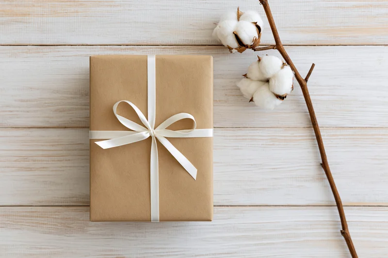 Kraft paper gift box tied with white satin ribbon on wooden surface beside cotton branch