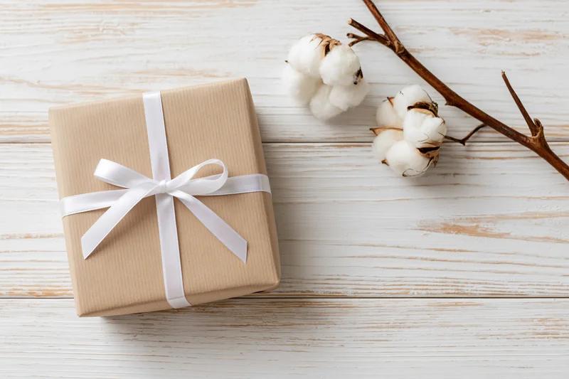 Kraft paper gift box tied with white satin ribbon on wooden surface beside cotton branch