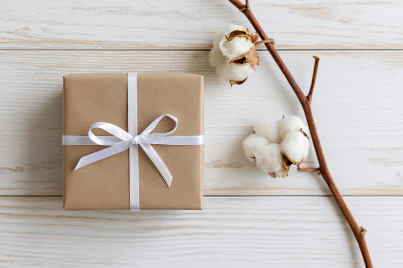Kraft paper gift box tied with white satin ribbon on wooden surface with cotton branch beside it